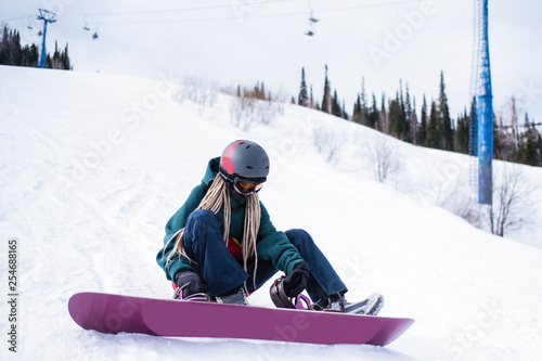 Woman snowboarder zipping up his boots while sitting on a snowy slope.