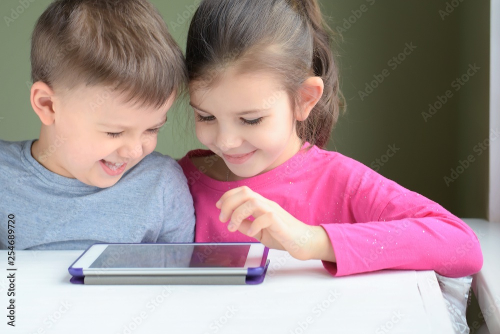 White caucasian toddler boy and beautiful girl playing together on tablet games. Portrait of smiling little brother and sister. Children play a computer game on the tablet White caucasian toddler boy and beautiful girl playing together on tablet games. Portrait of smiling little brother and sister. Children play a computer game on the tablet