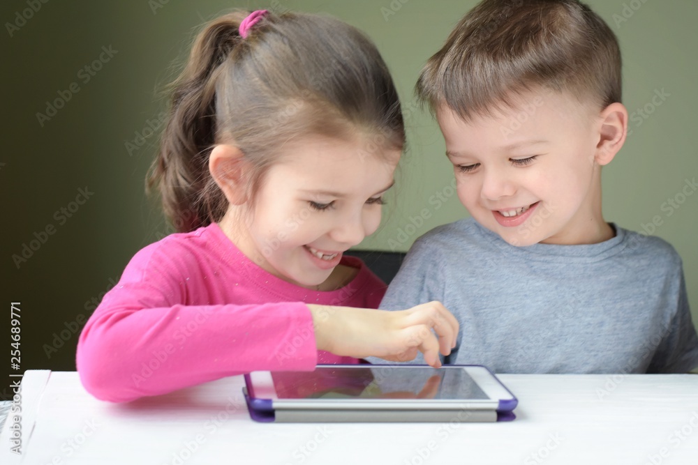 White caucasian toddler boy and beautiful girl playing together on tablet games. Portrait of smiling little brother and sister. Children play a computer game on the tablet White caucasian toddler boy and beautiful girl playing together on tablet games. Portrait of smiling little brother and sister. Children play a computer game on the tablet