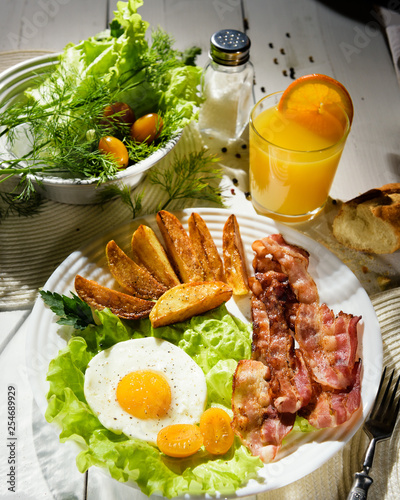 Breakfast with bacon. French fries and egg with bacon on top in a white bowl, on a large table with greens.