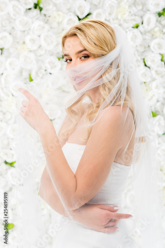 attractive young bride hiding face behind bridal veil while posing at camera on white floral background