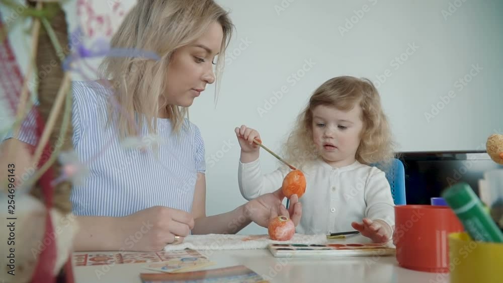 Happy Easter! Mother and her little daughter painting Easter eggs at the festive table with basket and eggs. Slow Motion