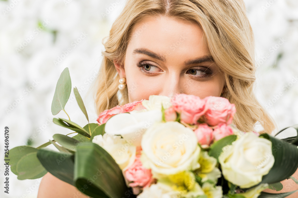 selective focus of pretty young bride enjoying flavor of wedding bouquet on white floral background