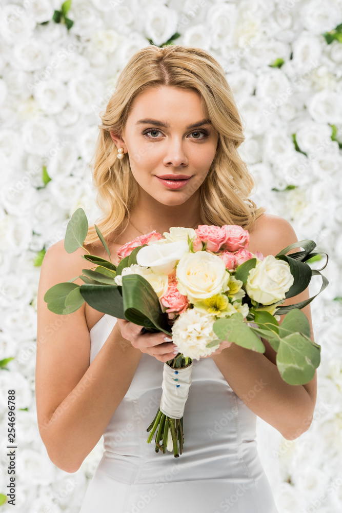 Naklejka premium beautiful young woman holding wedding bouquet and looking at camera on white floral background
