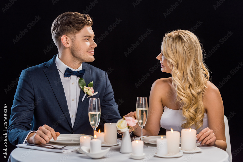 happy young couple sitting at served table and looking at each other isolated on black