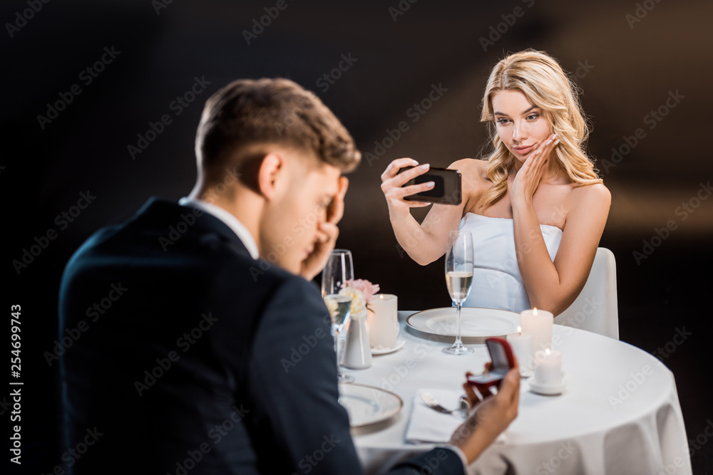 selective focus of pretty young woman taking selfie with smartphone, while sad man holding gift box with wedding ring on black background