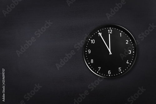 Black wall clock showing five minutes to midnight on black chalkboard background. Office clock showing 5 min to midnight or midday on black texture
