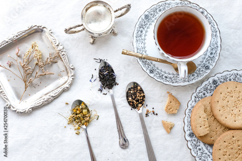 Different types of tea on vintage metal spoons on white fabric background, teacup, dried flower, silver plate, cookies. Vintage food and drink setting styling.  Organic healthy well-being lifestyle.