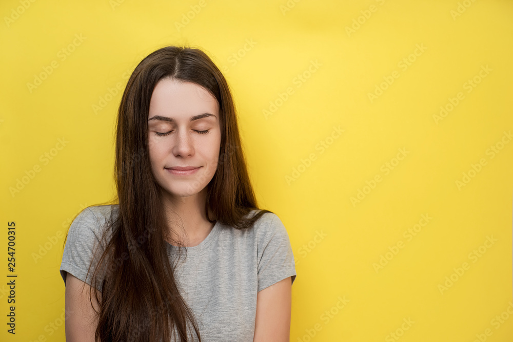 Close up shot of pleased adorable young female with wears gray T-shirt, has soap smile, keeps eyes closed, dreams about something pleasant. Attractive woman poses indoor on yellow will Close up shot of pleased adorable young female with wears gray T-shirt, has soap smile, keeps eyes closed, dreams about something pleasant. Attractive woman poses indoor on yellow will