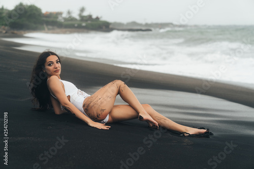 Charming girl with beautiful body lies on black sand beach. Sexy woman rests on summer vacation near ocean.