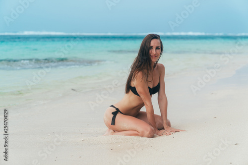 Sexy girl with sporty body in black bikini poses on beach with blue ocean at background.