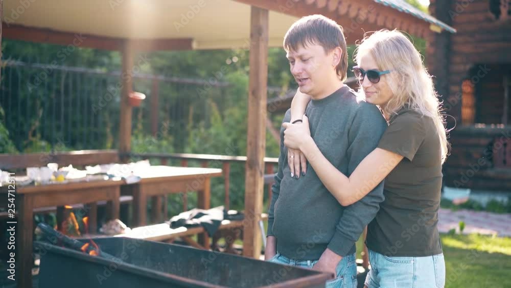 Travelling: Young guy with girl, standing near the barbecue, in which coals are smoking, against the backdrop of a summer house, on vacation. They talk and smile. Against the background of the sunset. Travelling: Young guy with girl, standing near the barbecue, in which coals are smoking, against the backdrop of a summer house, on vacation. They talk and smile. Against the background of the sunset.