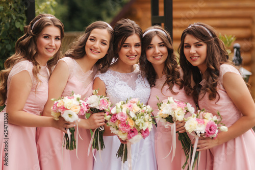 Happy bride with bridesmaid hold bouquets and have fun outside. Beautiful bridesmaid in same dresses stand by the charming bride in long wedding dress
