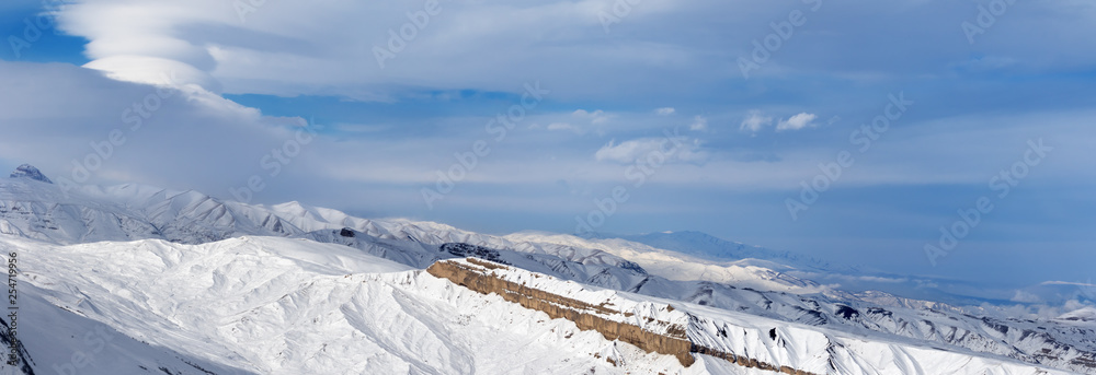Panorama of snowy winter mountains and cloudy blue sky at sun evening