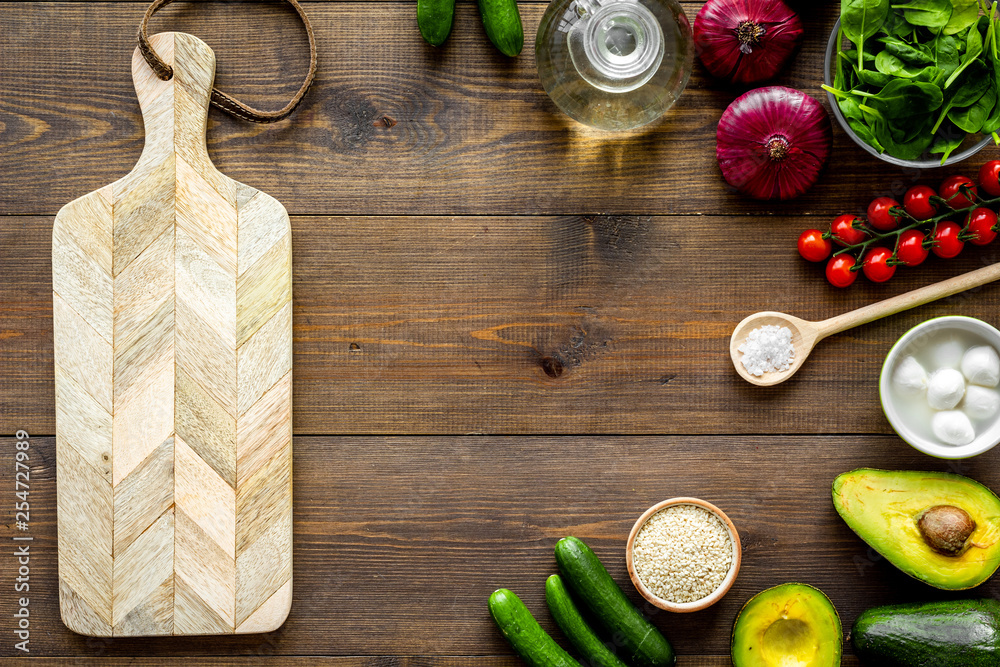 Ingredients for fresh vegetable salad and cutting board on dark wooden kitchen desk top view copy space mockup frame