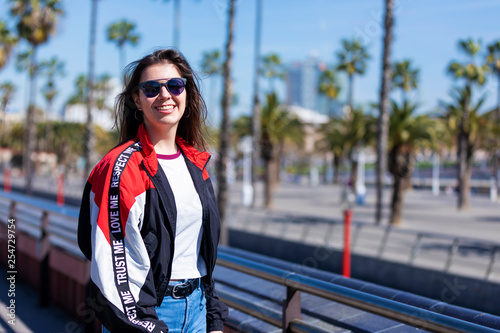 Front view of beautiful smiling young woman wearing urban clothes and sunglasses standing while looking camera outdoors in the street in a bright day