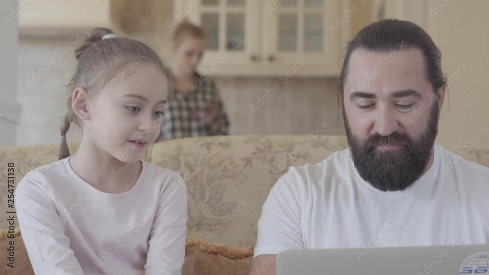 Bearded man sitting on the sofa and talking to his little daughter close up. Concept of happy family. Small girl sharing her secrets with dad sitting near laptop Bearded man sitting on the sofa and talking to his little daughter close up. Concept of happy family. Small girl sharing her secrets with dad sitting near laptop