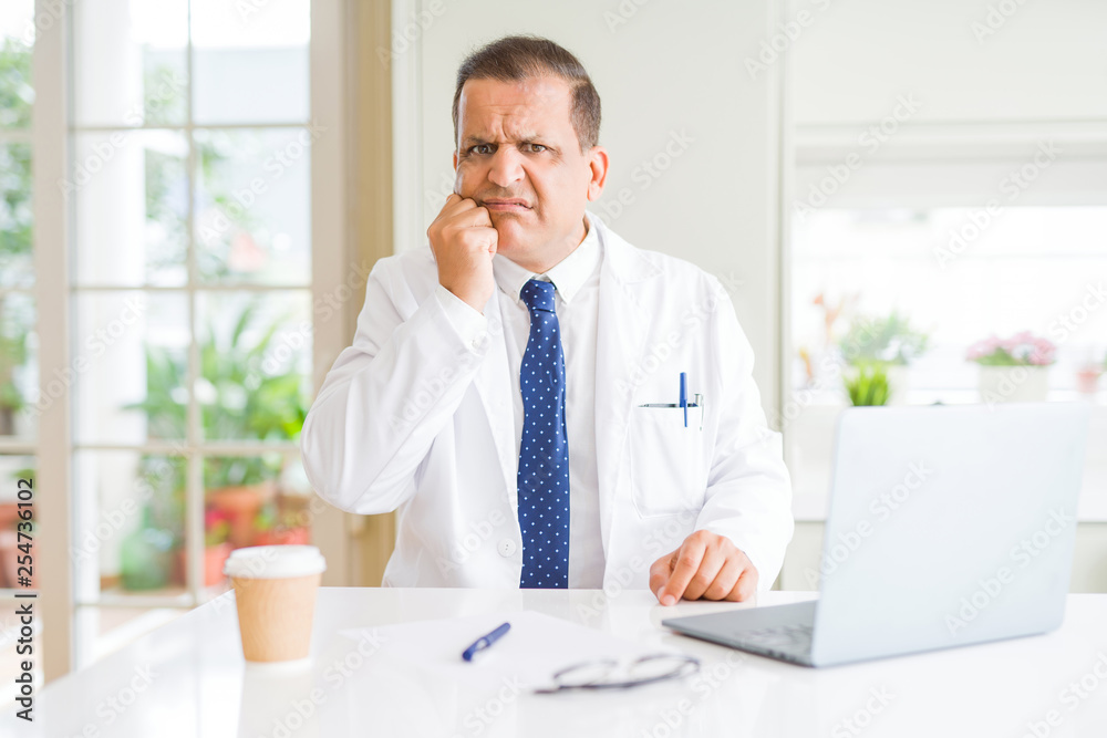 Middle age doctor man wearing white medical coat working with laptop at the clinic looking stressed and nervous with hands on mouth biting nails. Anxiety problem.