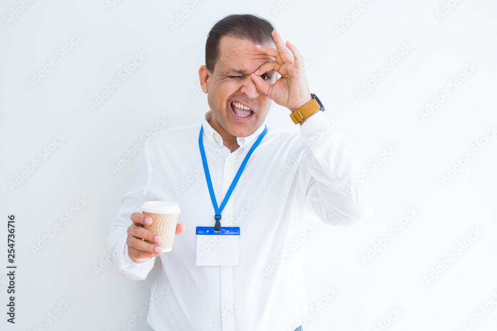 Middle age business man wearing ID card and drinking coffeeover white background with happy face smiling doing ok sign with hand on eye looking through fingers Middle age business man wearing ID card and drinking coffeeover white background with happy face smiling doing ok sign with hand on eye looking through fingers