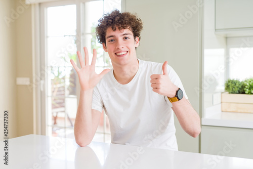 Young handsome man wearing white t-shirt at home showing and pointing up with fingers number six while smiling confident and happy.
