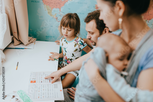 Mother holds a baby while father helps his little daughter to make a lessons at the desk in the room with the map on the wall
