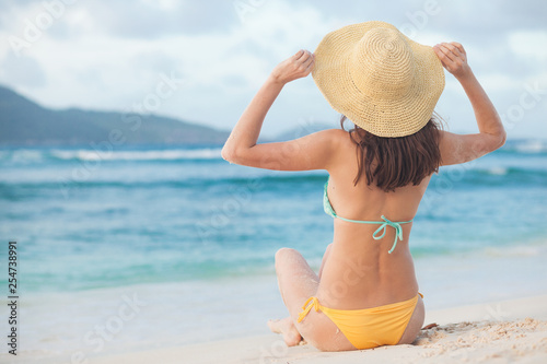 back view of long haired woman in bikini and straw hat relaxing on tropical beach. La Digue, Seychelles © el.rudakova