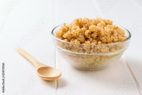 Glass bowl with boiled quinoa and wooden spoon on white table. A gluten-free boiled cereal dish.