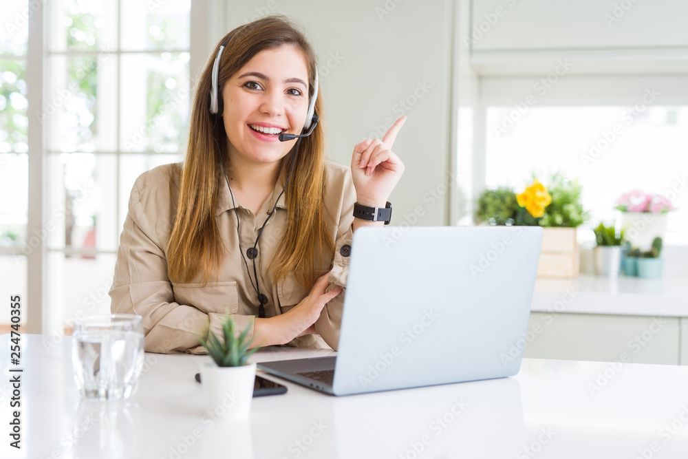 Beautiful young operator woman working with laptop and wearing headseat with a big smile on face, pointing with hand and finger to the side looking at the camera. Beautiful young operator woman working with laptop and wearing headseat with a big smile on face, pointing with hand and finger to the side looking at the camera.