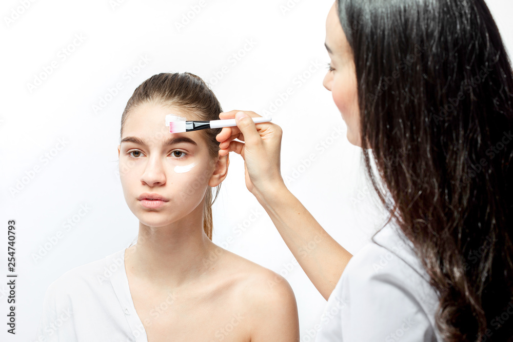 Cosmetologist applies with a brush white cosmetic mask on the face of young brown-haired girl