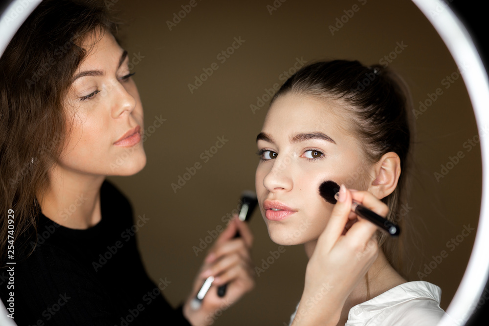 Reflection in the mirror of makeup artist the charming girl is doing makeup to a beautiful young girl Reflection in the mirror of makeup artist the charming girl is doing makeup to a beautiful young girl