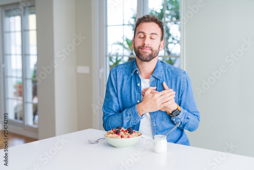 Handsome man eating cereals for breakfast at home smiling with hands on chest with closed eyes and grateful gesture on face. Health concept.