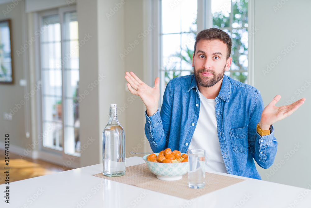Handsome man eating pasta with meatballs and tomato sauce at home clueless and confused expression with arms and hands raised. Doubt concept. Handsome man eating pasta with meatballs and tomato sauce at home clueless and confused expression with arms and hands raised. Doubt concept.