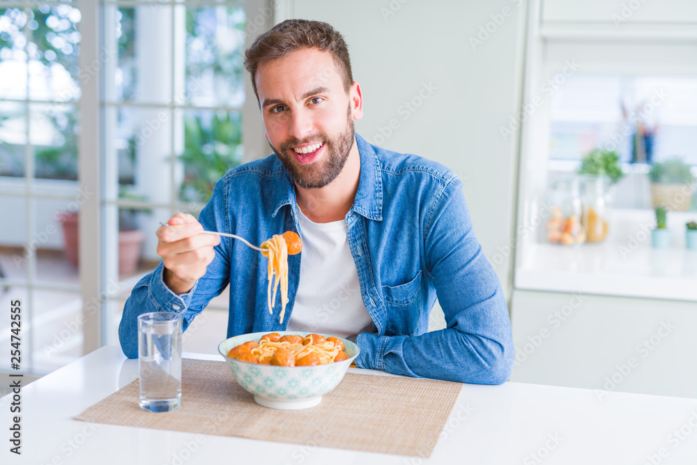 Handsome man eating pasta with meatballs and tomato sauce at home while smiling at the camera Handsome man eating pasta with meatballs and tomato sauce at home while smiling at the camera