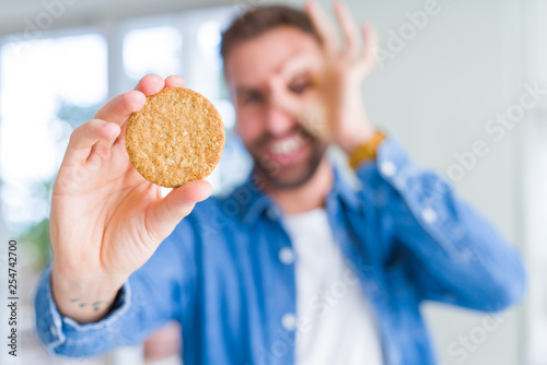 Handsome man eating healthy whole grain biscuit with happy face smiling doing ok sign with hand on eye looking through fingers