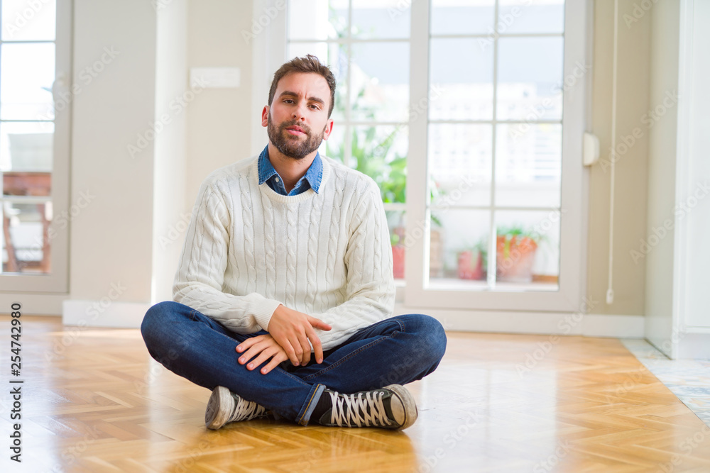 Handsome man wearing casual sweater sitting on the floor at home with serious expression on face. Simple and natural looking at the camera.