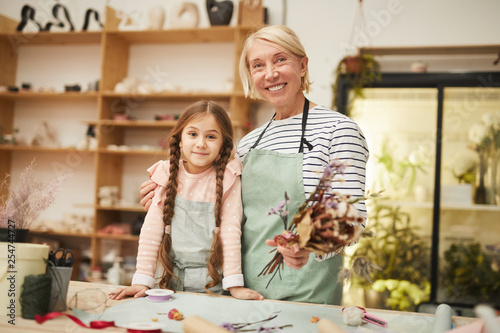 Waist up portrait of smiling mature woman looking at camera while creating flower compositions with cute girl in art studio, copy space