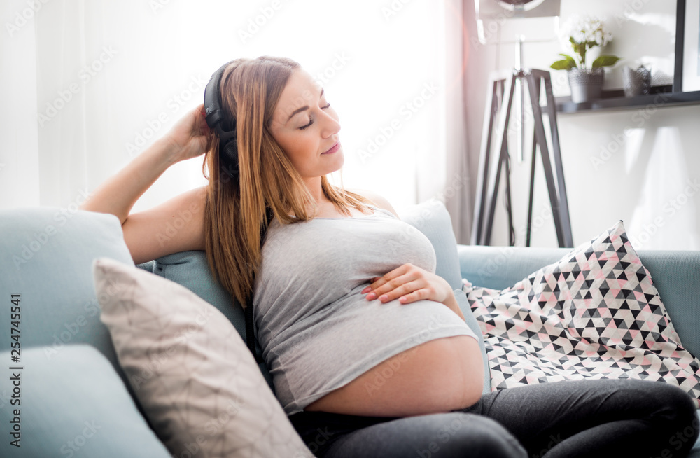 Relaxed pregnant woman sitting on sofa at home and listening music in headphones Relaxed pregnant woman sitting on sofa at home and listening music in headphones
