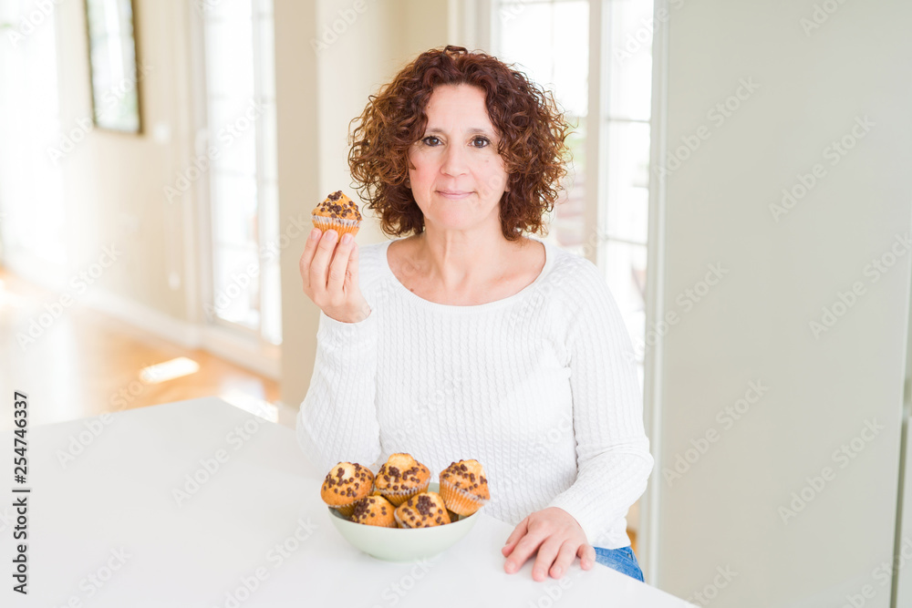Senior woman eating chocolate chips muffins with a confident expression on smart face thinking serious Senior woman eating chocolate chips muffins with a confident expression on smart face thinking serious