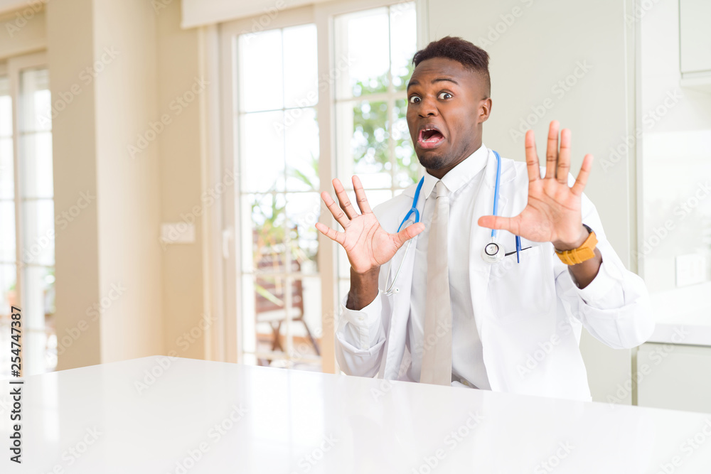 African american doctor man at the clinic afraid and terrified with fear expression stop gesture with hands, shouting in shock. Panic concept.