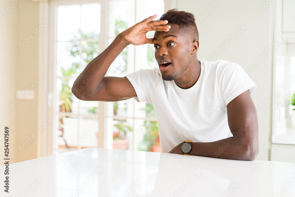 Handsome african american man on white table at home very happy and smiling looking far away with hand over head. Searching concept. Handsome african american man on white table at home very happy and smiling looking far away with hand over head. Searching concept.
