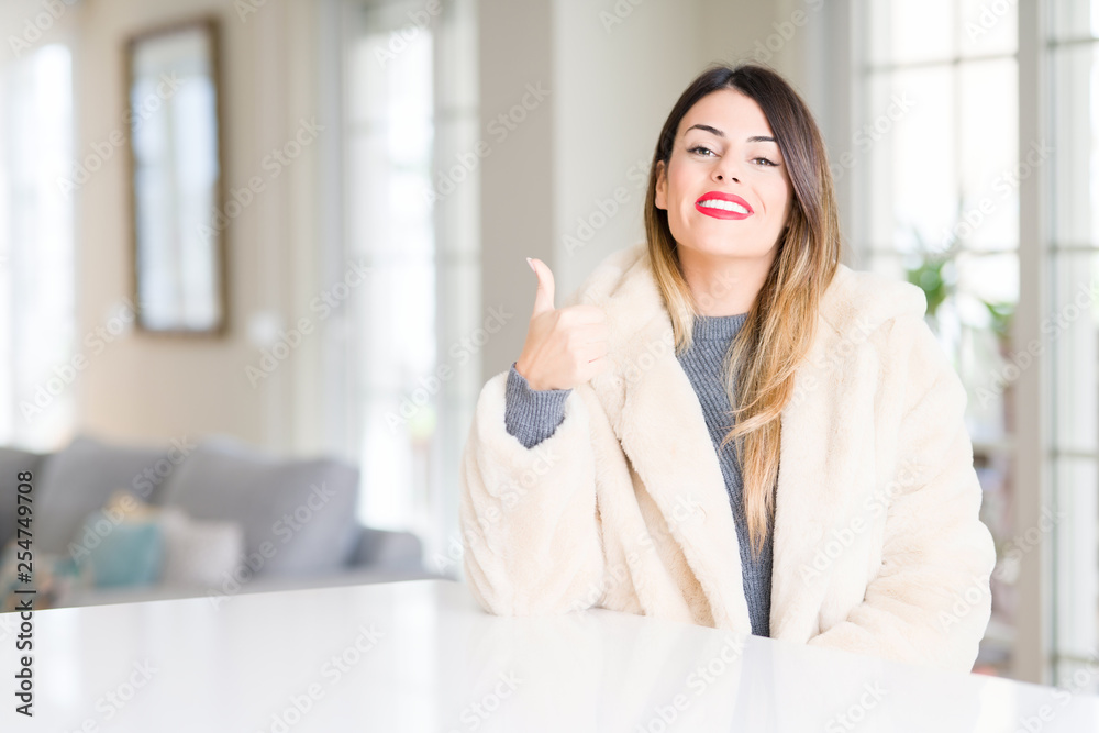 Young beautiful woman wearing winter fur coat at home doing happy thumbs up gesture with hand. Approving expression looking at the camera with showing success. Young beautiful woman wearing winter fur coat at home doing happy thumbs up gesture with hand. Approving expression looking at the camera with showing success.