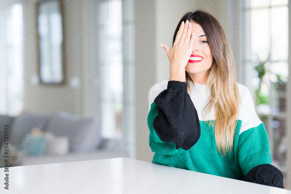 Young beautiful woman wearing winter sweater at home covering one eye with hand with confident smile on face and surprise emotion. Young beautiful woman wearing winter sweater at home covering one eye with hand with confident smile on face and surprise emotion.