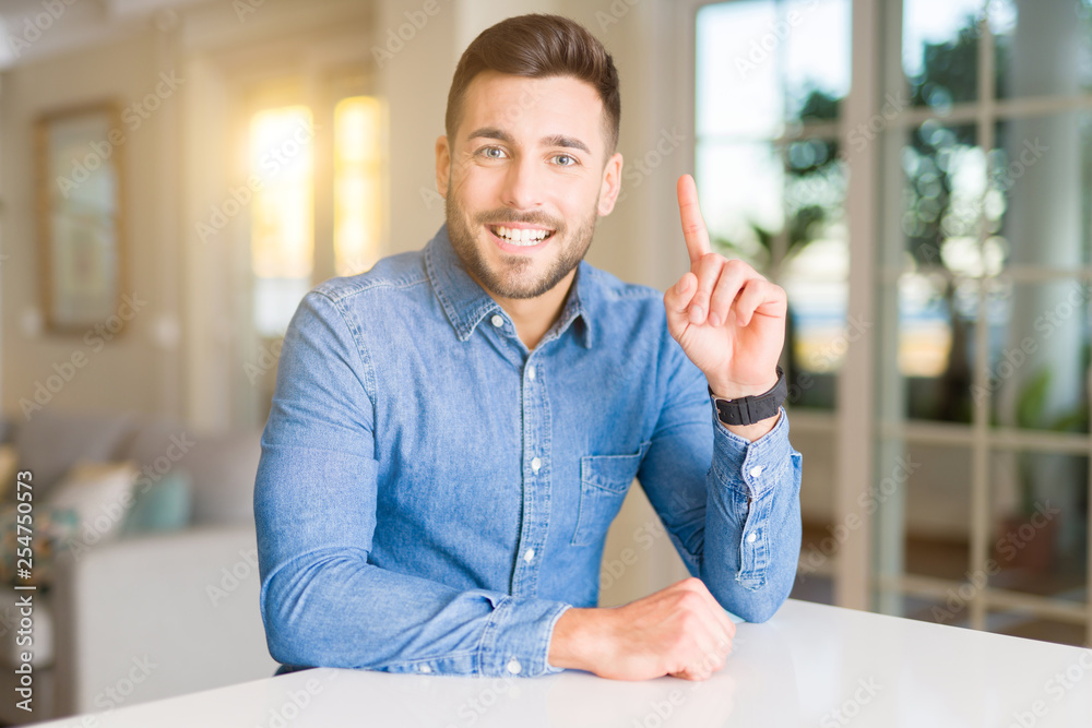 Naklejka premium Young handsome man at home surprised with an idea or question pointing finger with happy face, number one