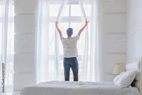 Young man near window at modern home. Man opening curtains in abstract white bedroom. Rear view photo