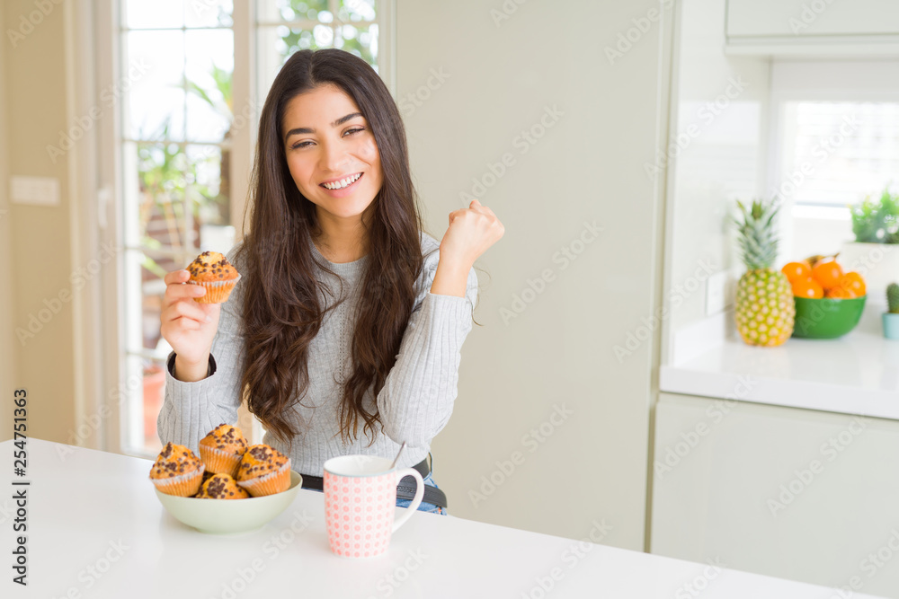 Young woman eating chocolate chips muffins and drinking coffee screaming proud and celebrating victory and success very excited, cheering emotion Young woman eating chocolate chips muffins and drinking coffee screaming proud and celebrating victory and success very excited, cheering emotion
