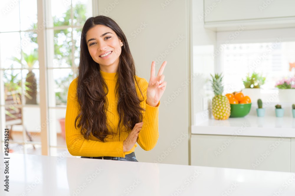 Young beautiful woman at home on white table smiling with happy face winking at the camera doing victory sign. Number two. Young beautiful woman at home on white table smiling with happy face winking at the camera doing victory sign. Number two.