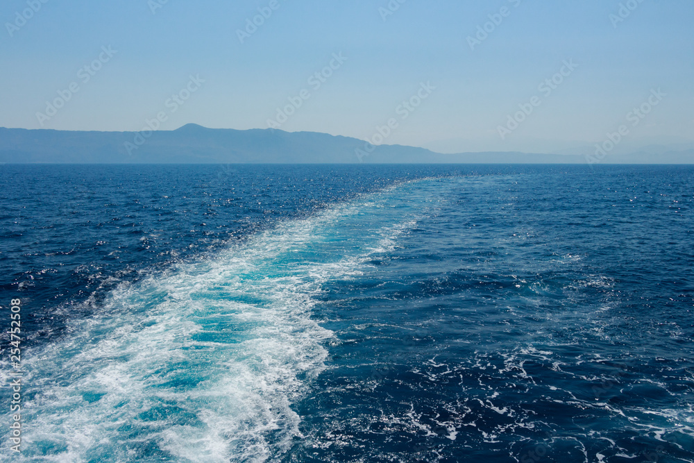 Island silhouette in the background. View from a boat with long path of waves from a boat. Deep dark blue water. Mediterranean Sea. Island silhouette in the background. View from a boat with long path of waves from a boat. Deep dark blue water. Mediterranean Sea.