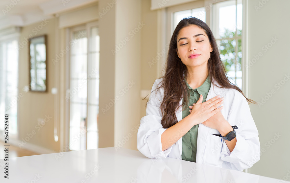 Young woman wearing medical coat at the clinic as therapist or doctor smiling with hands on chest with closed eyes and grateful gesture on face. Health concept. Young woman wearing medical coat at the clinic as therapist or doctor smiling with hands on chest with closed eyes and grateful gesture on face. Health concept.