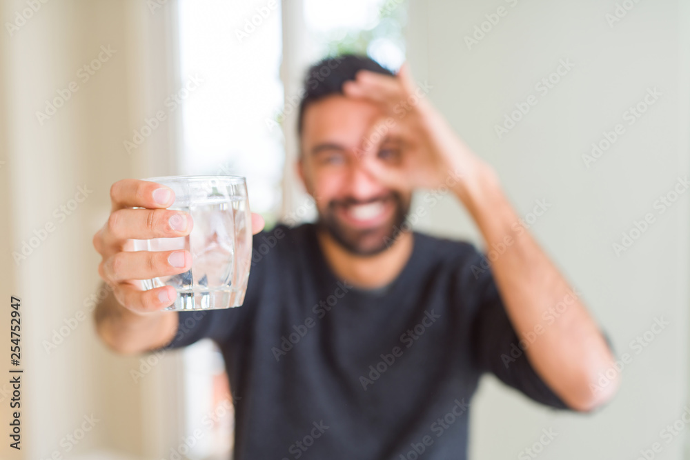 Handsome hispanic man drinking a fresh glass of water with happy face smiling doing ok sign with hand on eye looking through fingers Handsome hispanic man drinking a fresh glass of water with happy face smiling doing ok sign with hand on eye looking through fingers
