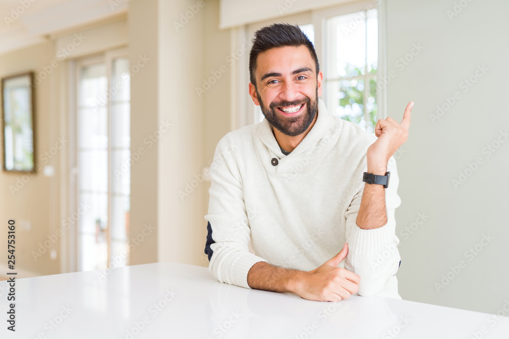 Handsome hispanic man wearing casual white sweater at home with a big smile on face, pointing with hand and finger to the side looking at the camera.